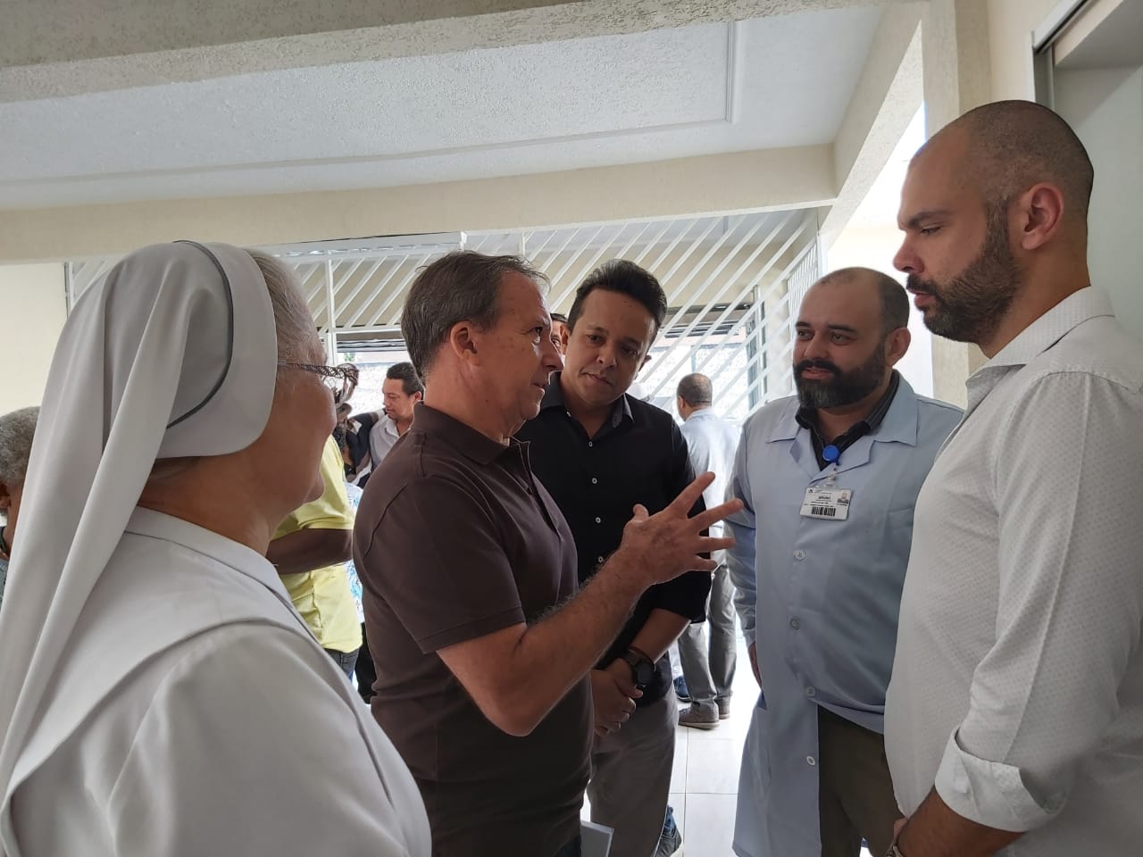 O prefeito, Bruno Covas, e o gerente da unidade do CAPS conversam com um homem branco de camiseta marrom. A conversa é acompanhada por um freira trajada de branco e pelo subprefeito, Gilmar Souza Santos. 